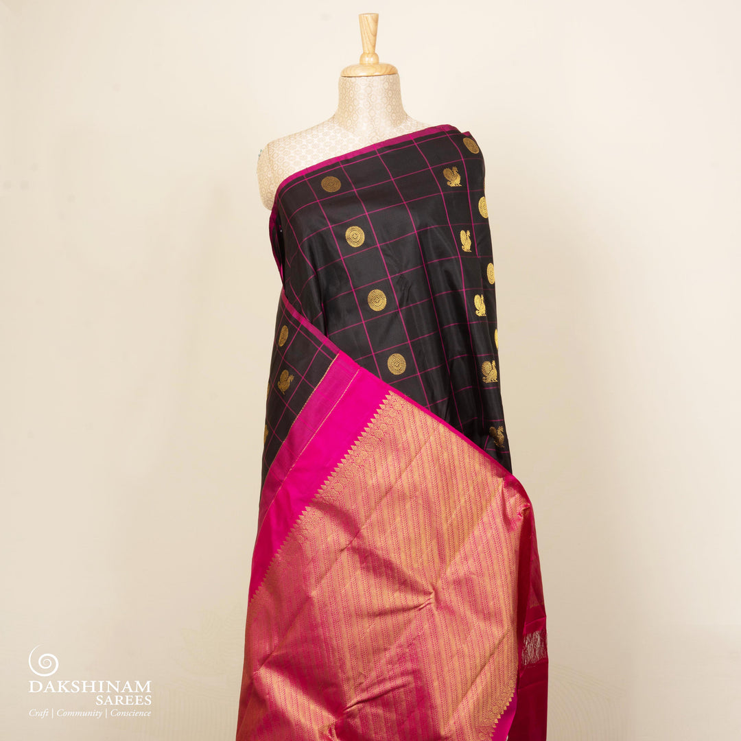 Black handwoven Kanjivaram silk saree with magenta checks, annam and chakram motifs, and pink pallu with gold zari chevron and leaf designs, paired with magenta blouse.