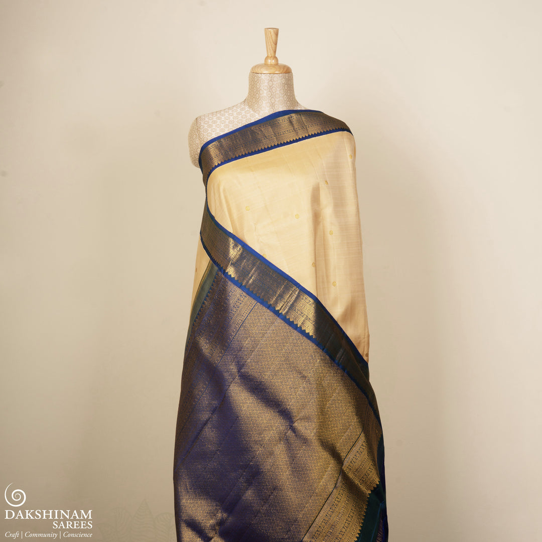 Cream Kanjivaram silk saree with chakram buttas, peacock blue border featuring chevron, rudraksham and floral lines, paired with plain blue blouse.