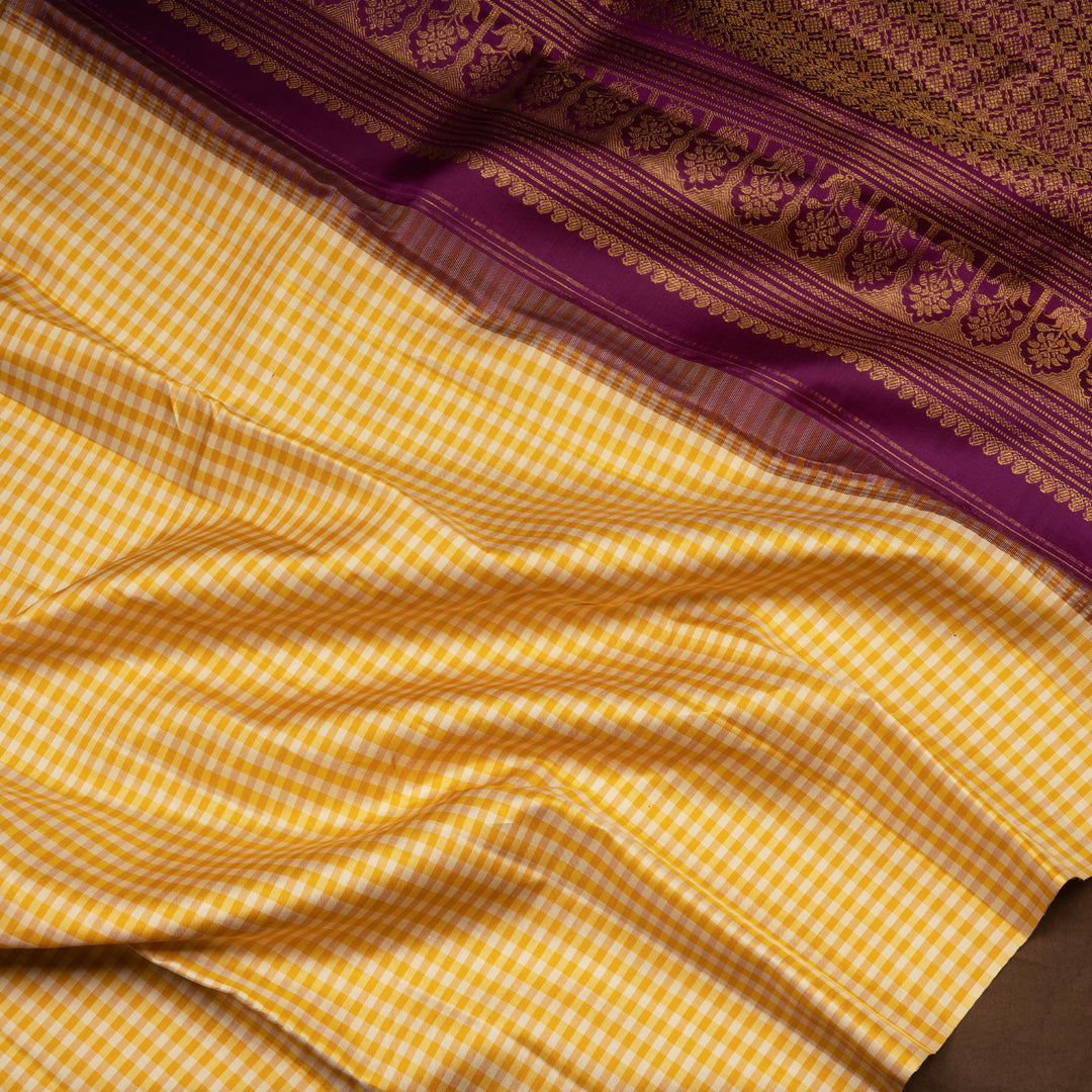 Beige and yellow small checked Kanjivaram silk saree with purple pallu featuring floral motifs, chevron and chakram designs, paired with dotted butta blouse.