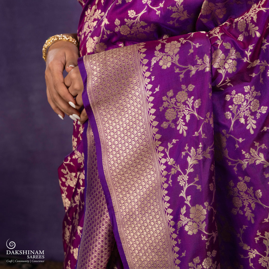 Magenta and violet mixed Banarasi silk saree with gold zari floral buttas, beige leaf panel border and matching beige leaf panel pallu, paired with a magenta-violet blouse with beige leaf border.