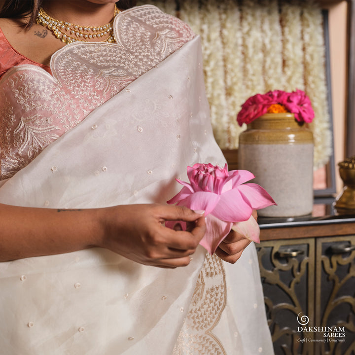 Person wearing a white saree with pink blouse, holding a pink flower, with a decorative background.